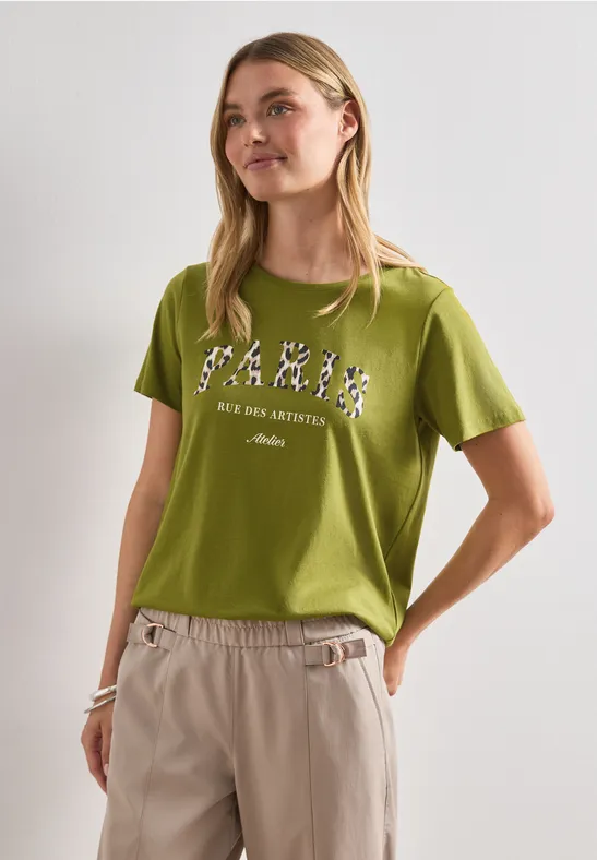 Person in grünem T-Shirt mit Print, beigen Hosen, seitlich vor weißem Hintergrund.