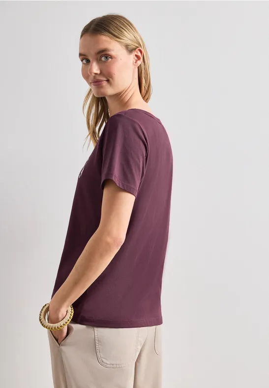 Seitlich stehende Person in lila T-Shirt und beigen Hosen, das Gesicht unkenntlich gemacht.