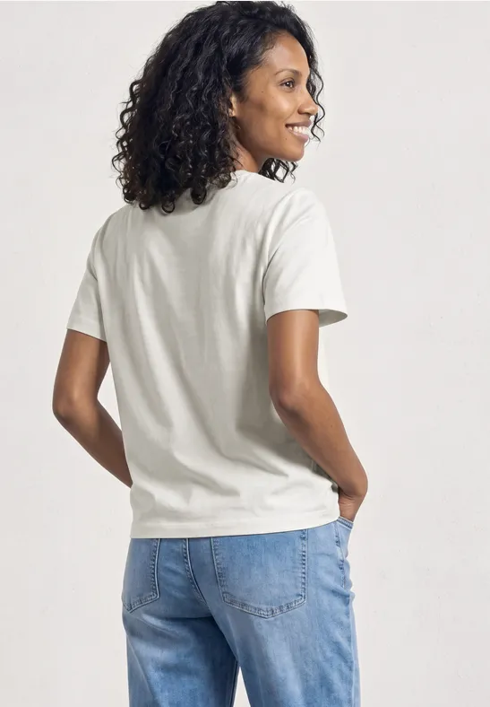 Een persoon staat met de rug naar de camera, draagt een wit T-shirt en blauwe jeans, met donker krullend haar.