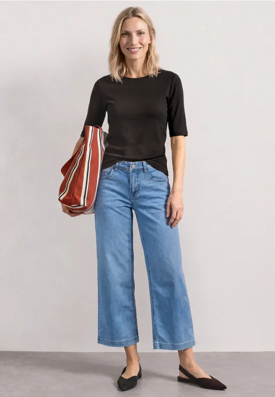 Vrouw draagt zwart shirt met korte mouwen, wijde blauwe jeans, zwarte ballerina’s en een gestreepte tas.