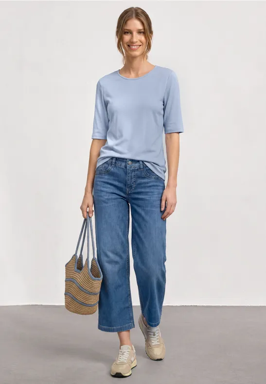 Vrouw met lichtblauw shirt, wijde cropped jeans, beige sneakers en een gestreepte strooien tas.