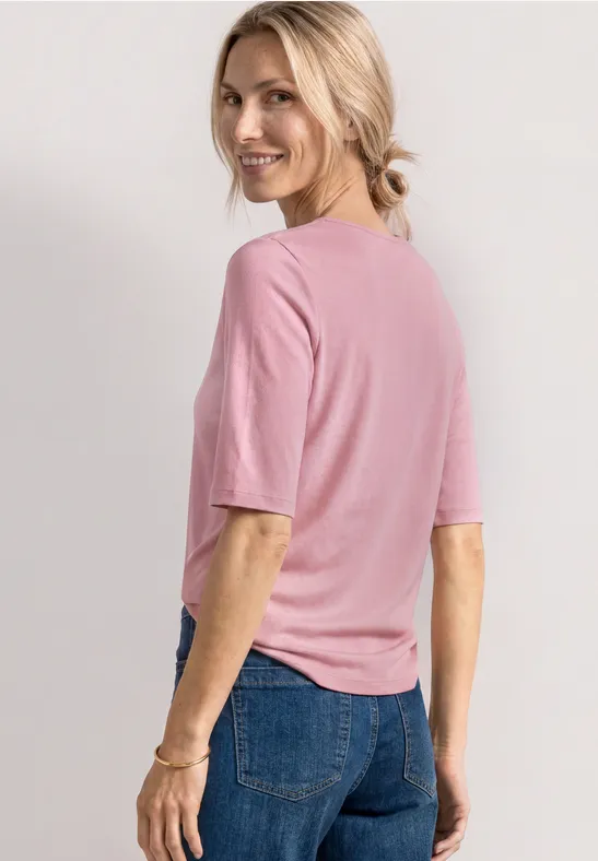 Rugaanzicht van een persoon in een roze shirt met halve mouw en blauwe jeans, blond haar in paardenstaart.