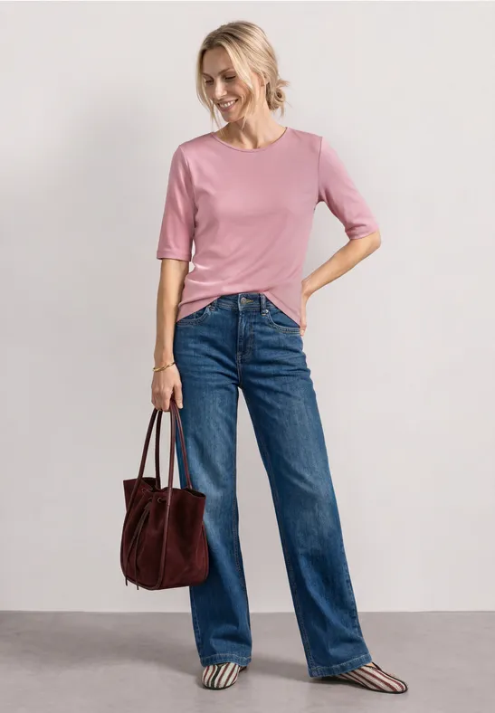 Een vrouw draagt een roze shirt, blauwe spijkerbroek en houdt een bruine handtas vast.