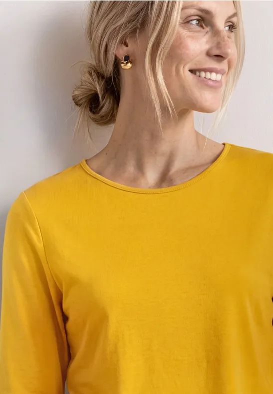 Persoon met gele top, gouden creolen en lage knot voor een lichte, neutrale achtergrond.
