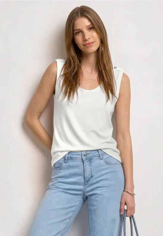 Eine Frau steht vor weißem Hintergrund, trägt ein weißes Top, blaue Jeans und hält ein Handy.