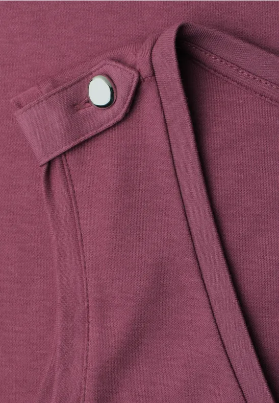 Nahaufnahme eines bordeauxfarbenen Shirts mit schmalem Riegel und silbernem Druckknopf am Ausschnitt.