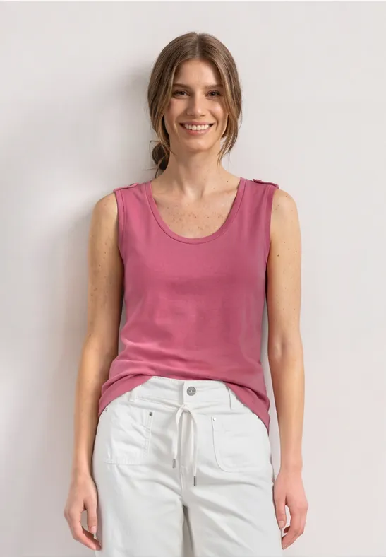 Eine Frau steht vor weißem Hintergrund, trägt rosa Top und weiße Hose.