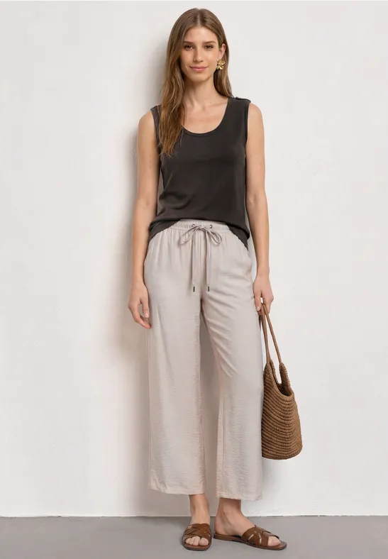 Frau trägt braunes ärmelloses Top, helle, weite Leinenhose, Basttasche und flache Sandalen.