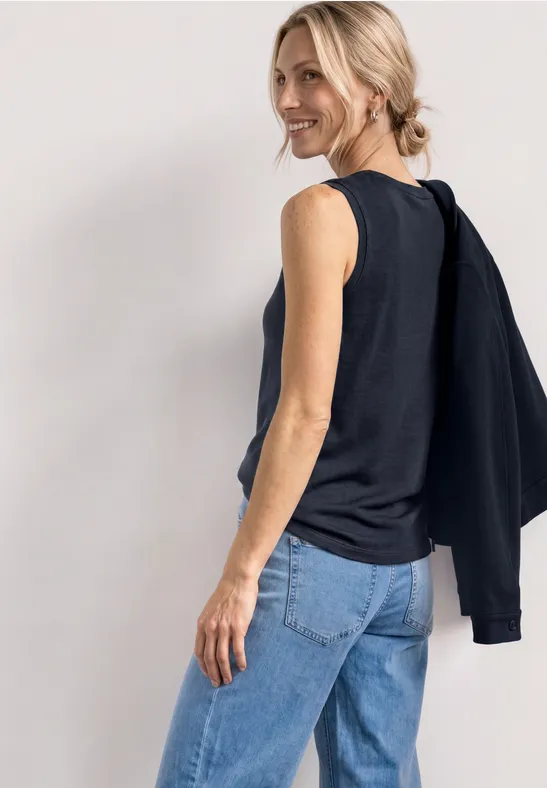 Frau seitlich in ärmellosem schwarzen Top und Jeans, mit schwarzem Pullover über der Schulter.