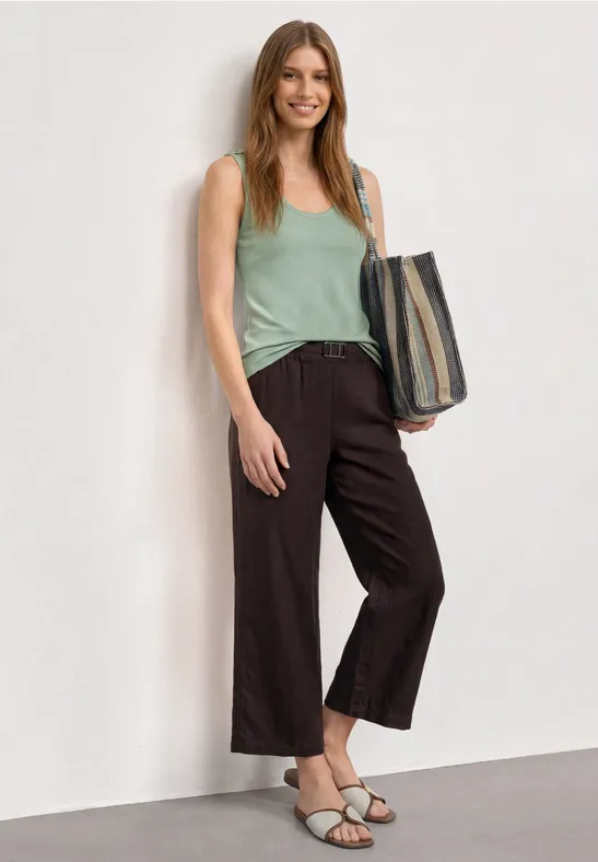 Femme en top vert et pantalon ample foncé, sac à main, sandales sur fond clair.