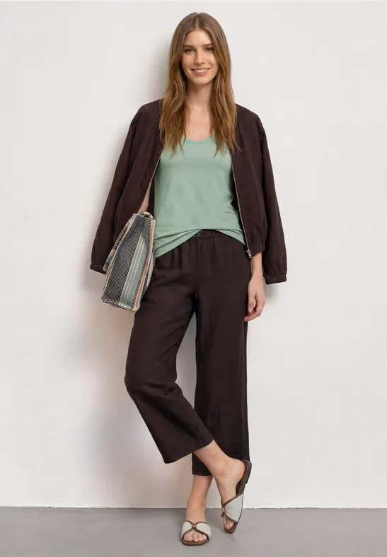 Une femme devant un mur blanc, vêtue d'une veste marron, pantalon ample, top vert menthe, sandales blanches et sac rayé.