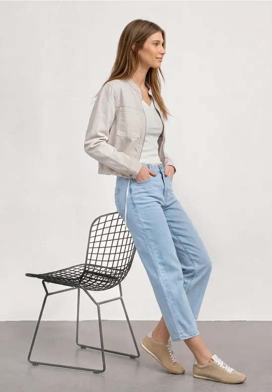 Vrouw leunt tegen gaasstoel, witte jas, wit shirt, lichtblauwe jeans en sneakers.