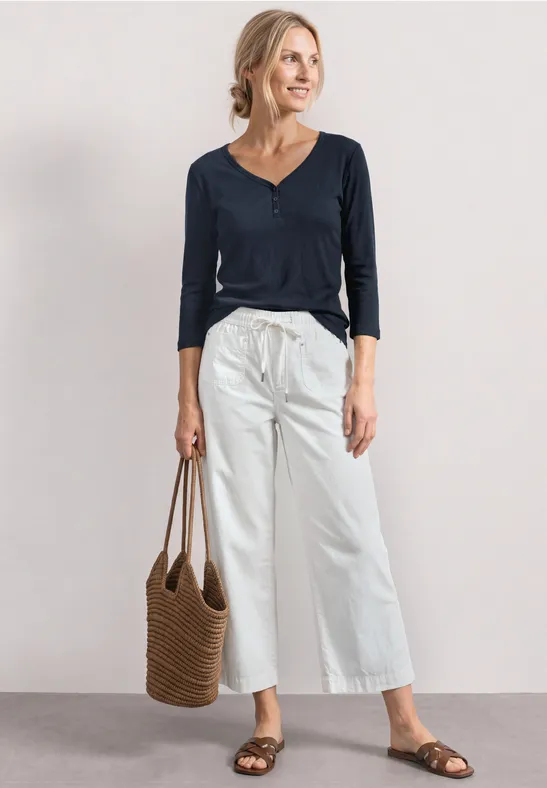 Vrouw in blauw shirt, wijde witte broek, bruine sandalen en strooien tas voor neutrale achtergrond.