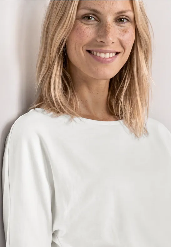 Blonde Frau mit schlichten weißen T-Shirt und mittellangen Haaren vor hellem Hintergrund.