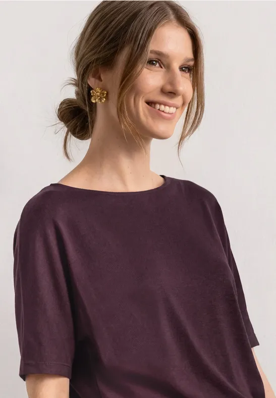 Frau mit hochgestecktem Haar trägt schlichtes lila Kurzarmshirt und goldenen Ohrschmuck.