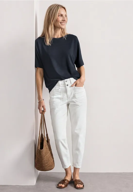 Vrouw tegen muur, zwart T-shirt, witte losse broek en gevlochten tas.