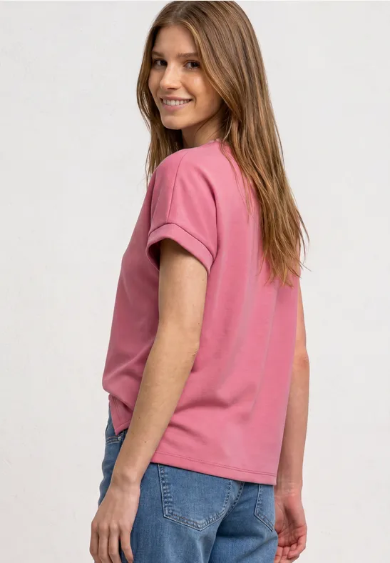 Eine junge Frau in rosa T-Shirt und blauen Jeans blickt lächelnd über die Schulter.