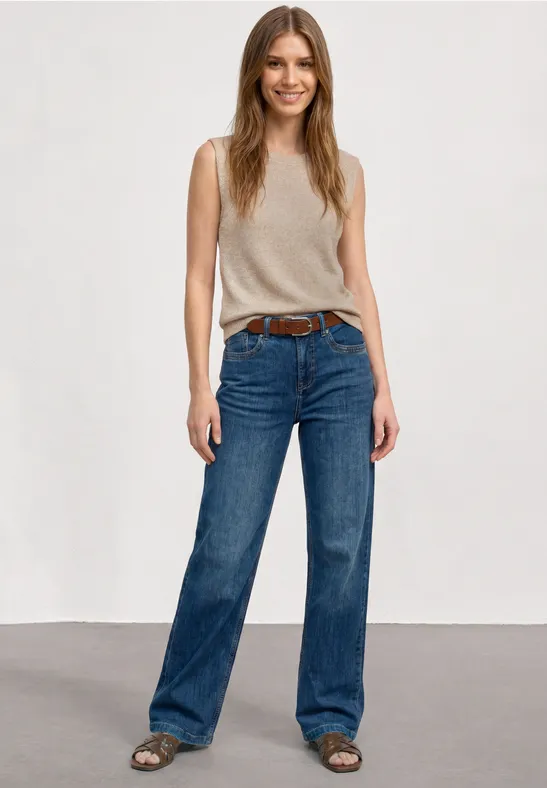 Frau mit langem Haar in beige ärmellosem Top, weiten blauen Jeans und Sandalen vor hellem Hintergrund.