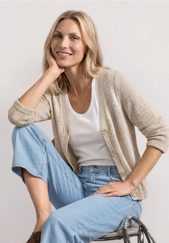 Eine Frau sitzt entspannt auf dem Boden, trägt helle Jeans, weißes Shirt und beigen Cardigan.