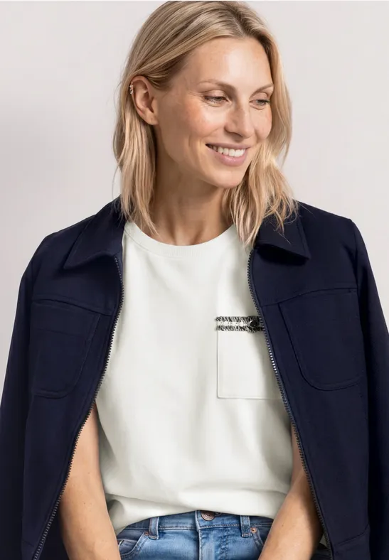 Vrouw met blond haar, draagt een wit t-shirt, blauwe jeans en een donker jasje over haar schouders.