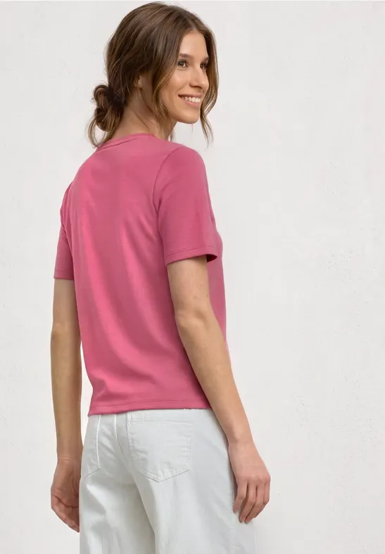 Rückansicht einer Frau in rosa T-Shirt und weißen Jeans vor neutralem Hintergrund.