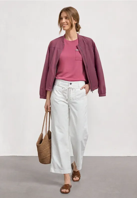 Eine Frau trägt rosa Shirt, violette Jacke, weiße Culotte, Sandalen und Korbtasche beim Gehen.