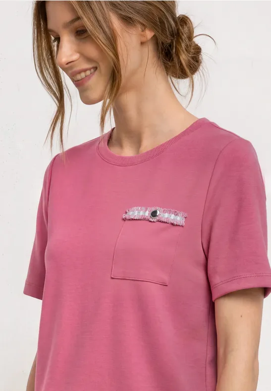 Eine junge Frau in einem rosafarbenen T-Shirt mit Brusttasche und Dekoration lächelt seitlich.