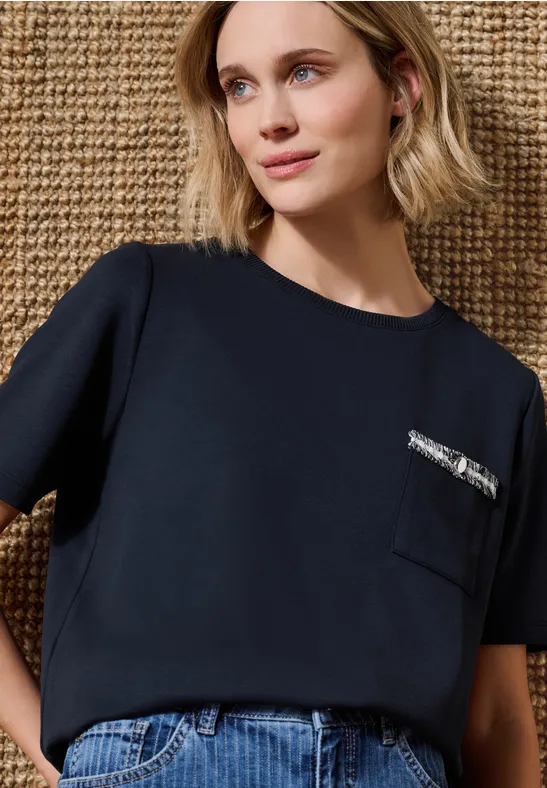 Eine Frau in dunkelblauem Shirt mit Brusttasche und Reißverschluss, Jeans vor hellem Naturhintergrund.