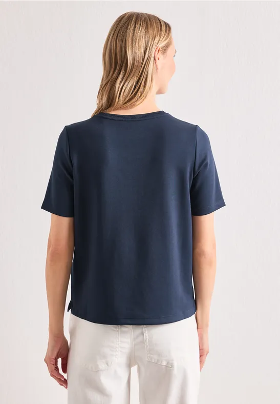 Frau von hinten in dunkelblauem T-Shirt und weißer Hose vor hellem Hintergrund.