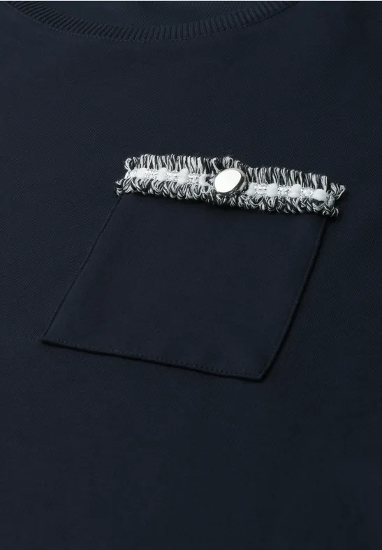 Marineblaues T-Shirt mit Brusttasche und darüber liegender Fransenborte, verziert mit einem Metallknopf.