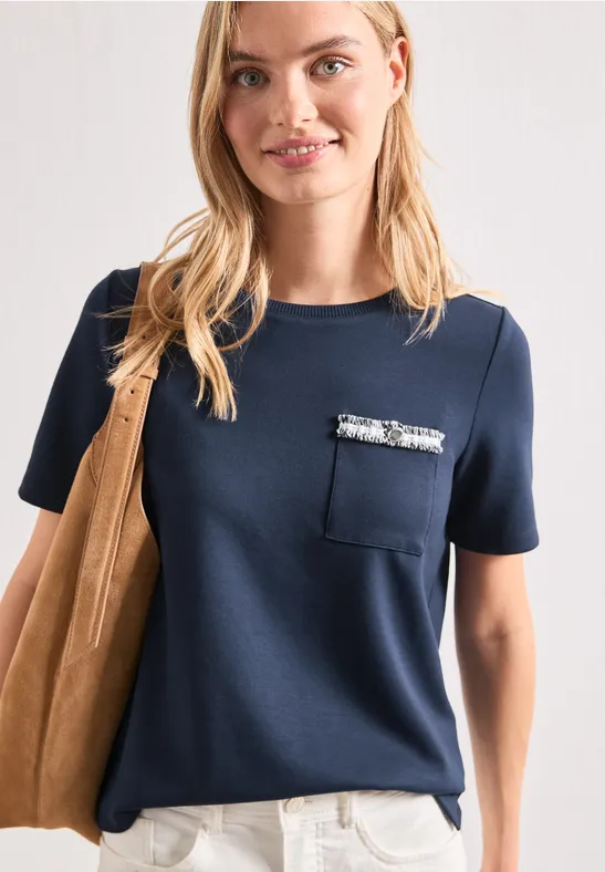 Frau in marineblauem, kurzärmeligem Shirt mit Brusttasche und Paillettenbesatz, weiße Hose und Wildleder-Schultertasche.
