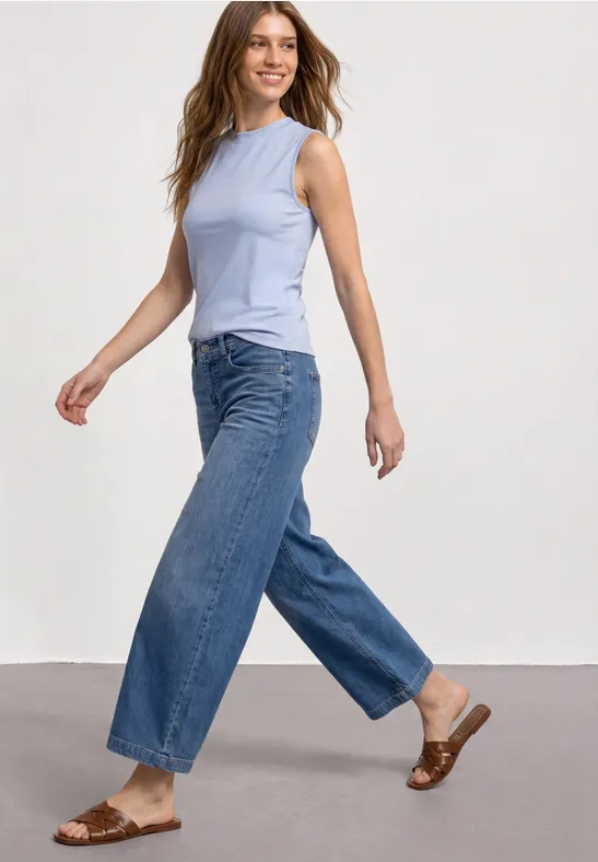 Vrouw in mouwloze lichtblauwe top, wijde jeans en bruine sandalen voor een neutrale achtergrond.