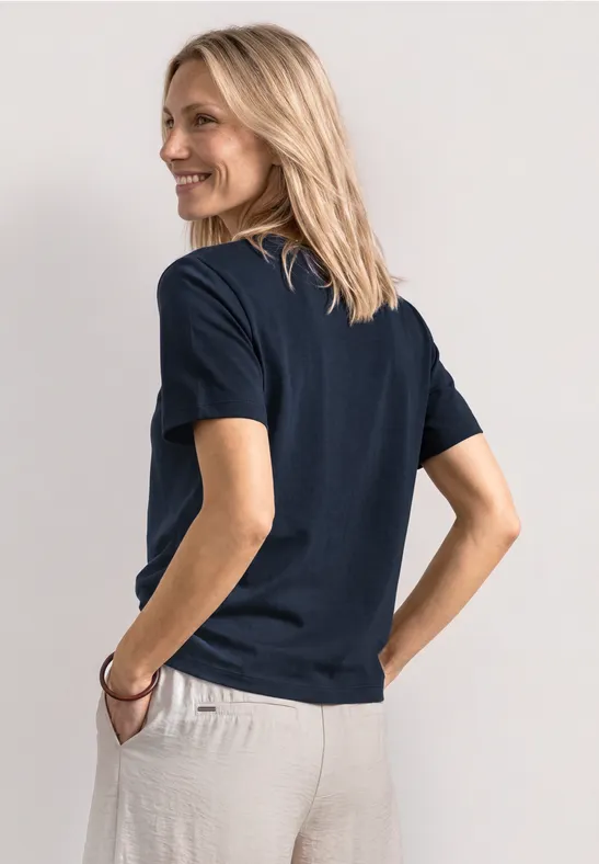 Rückansicht einer Person in dunkelblauem T-Shirt und hellbeiger Hose, Hand in Tasche.