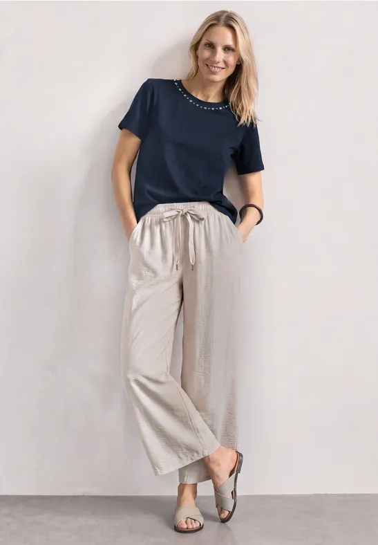 Frau in dunkelblauem T-Shirt, weiten beigen Hosen und Sandalen, lässig stehend mit einer Hand in der Hosentasche.