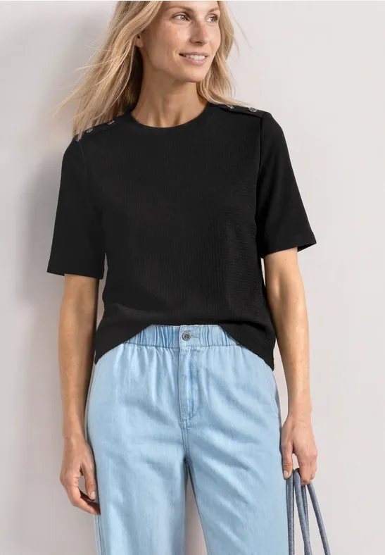 Frau trägt schlichtes schwarzes T-Shirt mit kurzen Ärmeln und hellblaue Jeans. Sie hält eine Tasche in der Hand.