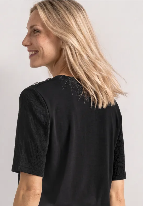 Eine Frau mit langen blonden Haaren, von hinten in schwarzem Kurzarmshirt vor hellem Hintergrund