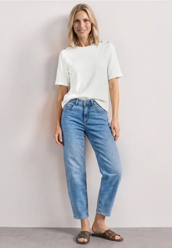 Frau trägt weißes T-Shirt, lockere blaue Jeans und braune Sandalen vor hellem Hintergrund.