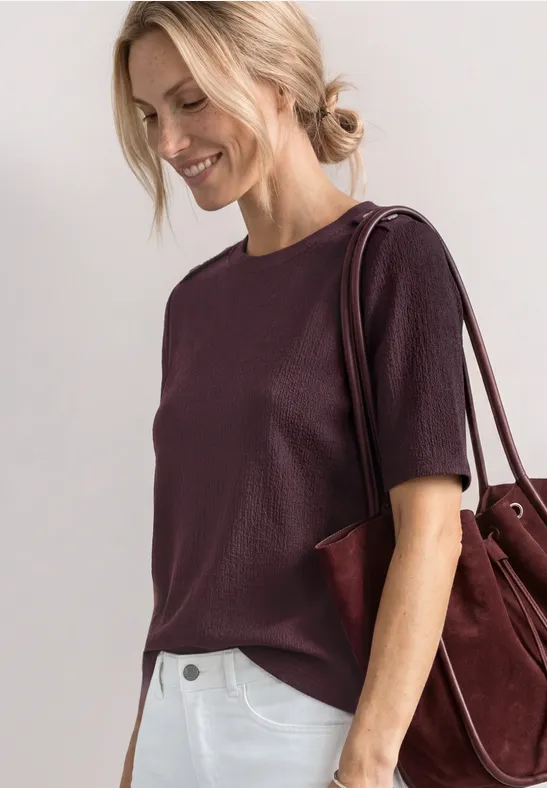 Frau in einem violetten Kurzarmshirt mit burgunderfarbener Schultertasche vor hellem Hintergrund.