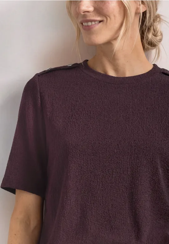 Nahaufnahme einer lächelnden Frau mit hochgesteckten blonden Haaren und bordeauxfarbenem Kurzarmshirt.