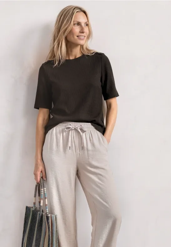 Frau vor weißer Wand in schwarzem T-Shirt, hellgestreifter Hose und schwarzer Tasche.