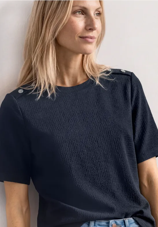 Frau trägt dunkelblaues, strukturiertes T-Shirt mit kurzen Ärmeln und dekorativen Knöpfen an den Schultern.
