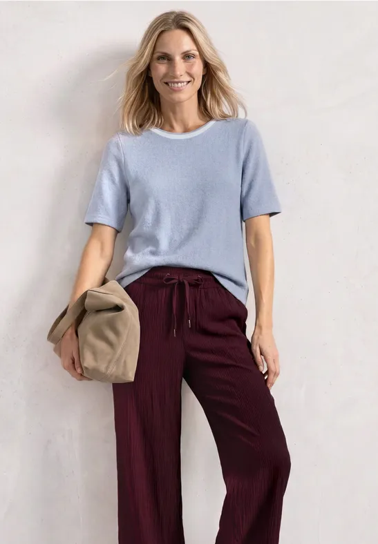 Vrouw met lichtblauw shirt en bordeauxrode broek, houdt een beige tas vast tegen een witte achtergrond.