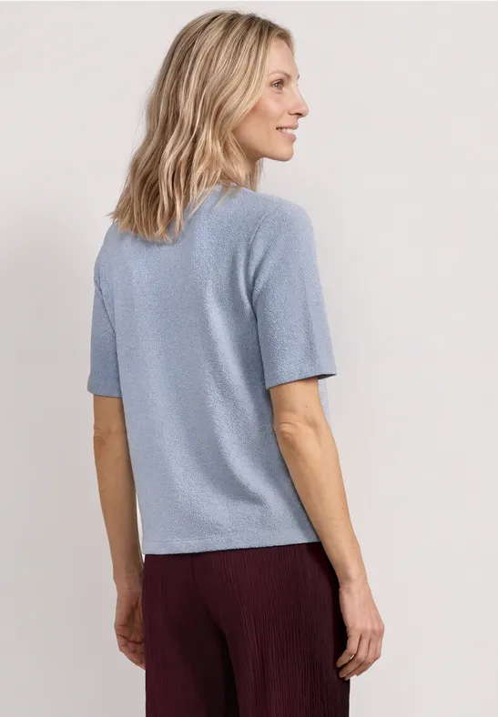 Vrouw van achteren in lichtblauw T-shirt met korte mouwen en bordeauxrode broek, halflang blond haar.