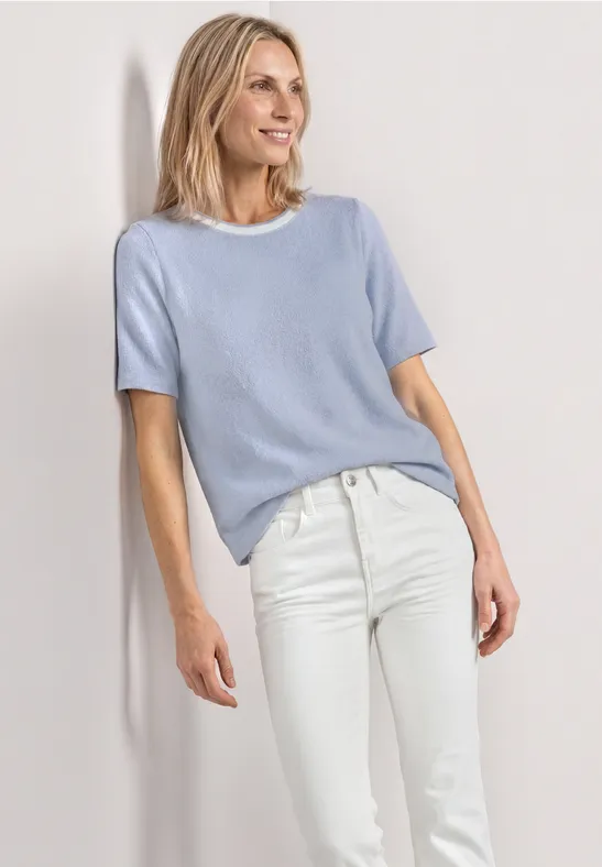 Frau in hellblauem Kurzarmpullover und weißen Jeans vor hellem Hintergrund