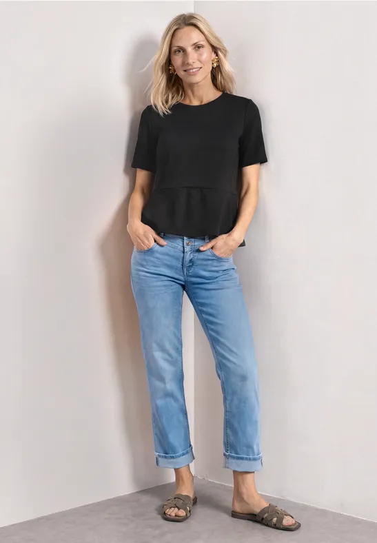 Vrouw in zwart T-shirt, blauwe jeans en sandalen, staand voor een lichte achtergrond.