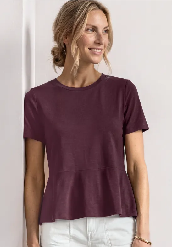 Frau trägt lila Kurzarmshirt mit ausgestelltem Saum und weiße Hose vor neutralem Hintergrund.