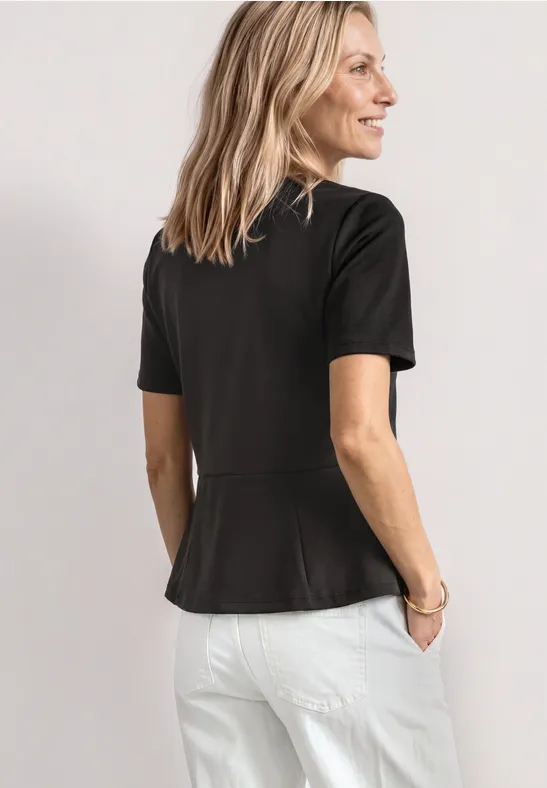 Een vrouw van achteren in een zwart T-shirt met korte mouwen en een witte broek voor een lichte achtergrond.