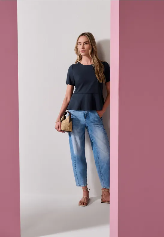 Een vrouw draagt een zwart peplumtop, lichtblauwe jeans en sandalen, met een handtas voor een roze achtergrond.