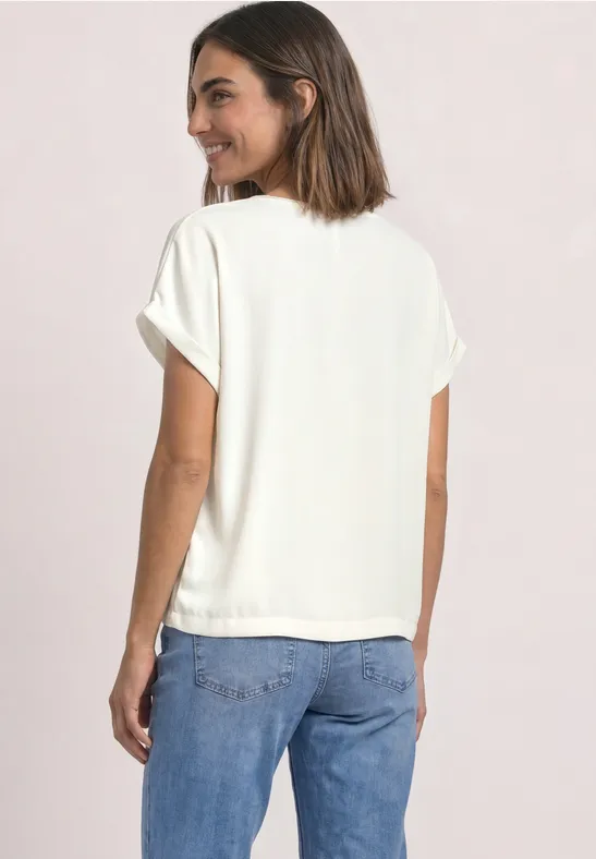 Eine Frau in weißem T-Shirt und blauen Jeans blickt über die Schulter und lächelt.