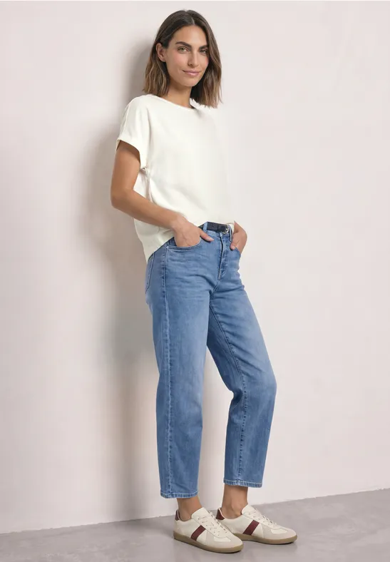 Eine Frau trägt ein weißes T-Shirt, blaue Jeans und weiße Sneakers und posiert vor hellem Hintergrund.